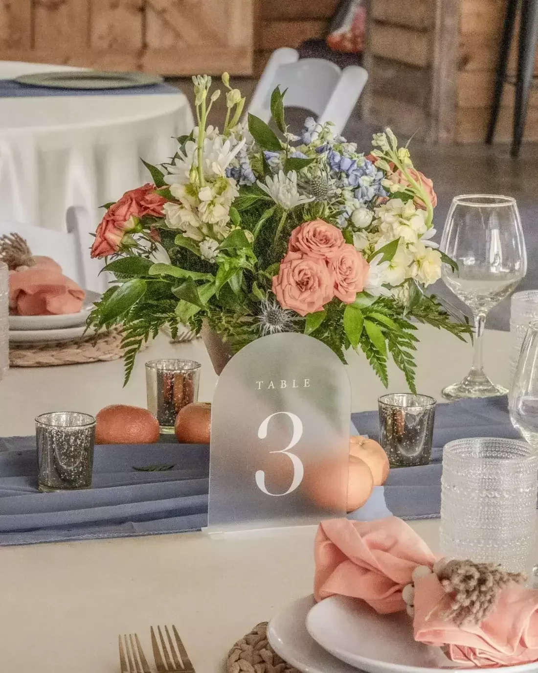 Wedding reception centerpiece of peach roses, blue hydrangea and greenery with a frosted table number